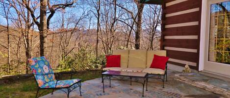 Un patio con un sofá y una silla, suelo de piedra y una cabaña de madera al fondo.