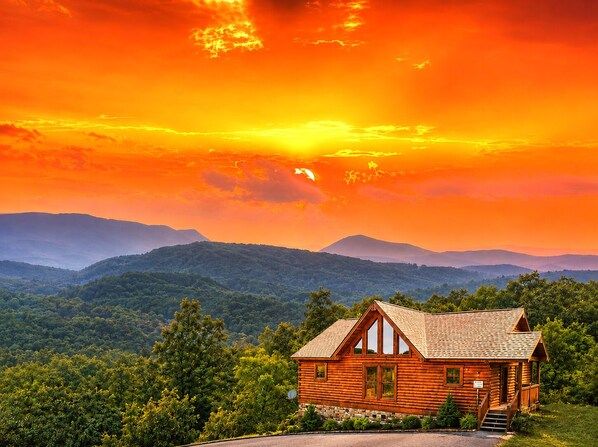 A wooden cabin with a stone foundation, surrounded by lush greenery and mountains in the background.