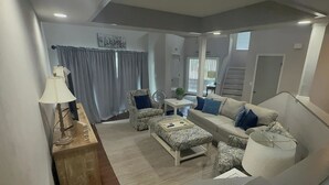 A living room with a sofa, ottoman, and a small table with a lamp. - AVAIL LABOR DAY WKEND - Steps to Beach - 3BR/3BA - Sleeps 8 - Garage (Palm Coast)