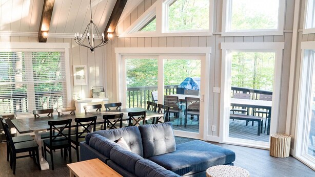 A spacious living area with a large blue sofa, wooden coffee table, and a wooden bench. There are multiple windows providing natural light and a view of the outdoors.