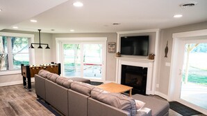 A modern living room with a sectional sofa, a foosball table, a fireplace, and a TV mounted above it.