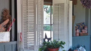 A rustic garden shed with open white shutters, a green potted plant, and various gardening tools.