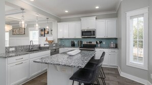 Una cocina moderna con armarios blancos, encimeras de granito, una isla central con taburetes y luces colgantes.