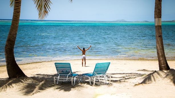 Une plage avec deux chaises longues bleues, une personne les bras levés, et une vue dégagée sur la mer turquoise.