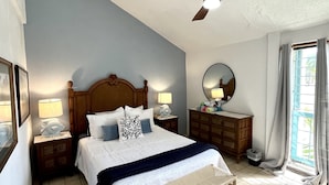 Une chambre à coucher comprenant un lit, des tables de chevet, un ventilateur de plafond, une fenêtre avec des rideaux et une vue sur la verdure à l’extérieur.