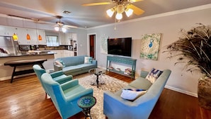 A living room with a sofa, armchairs, a coffee table, and a TV. There is a ceiling fan, a kitchen in the background, and a decorative plant.