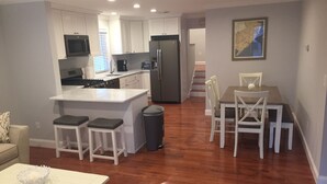 A modern kitchen with white cabinetry, a central island with bar stools, a dining area with a table and chairs, and a large painting on the wall. - Great Location / Walk to Everything!  4BR Big Backyard (Beach Haven)