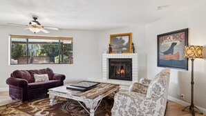A living room with a fireplace, a ceiling fan, a sofa, a patterned armchair, and a large window with a view of greenery. - Private Resort on 3/4 Acre Game Room Putting Green (Phoenix)