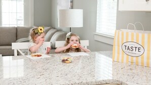 Twee kinderen zitten aan een keukentafel met eten, een lamp en een tas met het logo 'TASTE'.