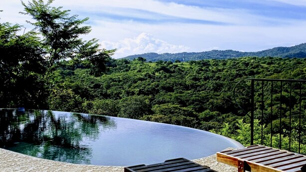 An infinity pool with wooden lounge chairs, surrounded by lush greenery and a mountainous backdrop. - AMAZING OCEAN VIEWS! "House in the Sky" Monte Paraiso (Playa Hermosa)