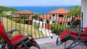 A balcony with two red chairs, a view of a coastal area with buildings and the sea. - AMAZING OCEAN VIEWS! "House in the Sky" Monte Paraiso (Playa Hermosa)