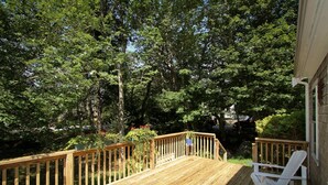A wooden deck with white chairs, surrounded by dense trees and a building on one side.