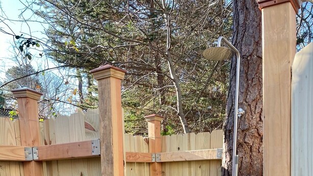 A wooden outdoor shower area with a metal showerhead, surrounded by a wooden fence, and a gravel ground.