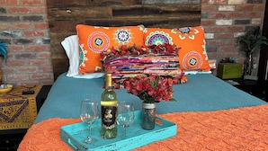 Un lit avec une couvre-lit aux couleurs turquoise et orange, un coussin décoratif, un vase avec des fleurs et un plateau avec une bouteille et des verres.
