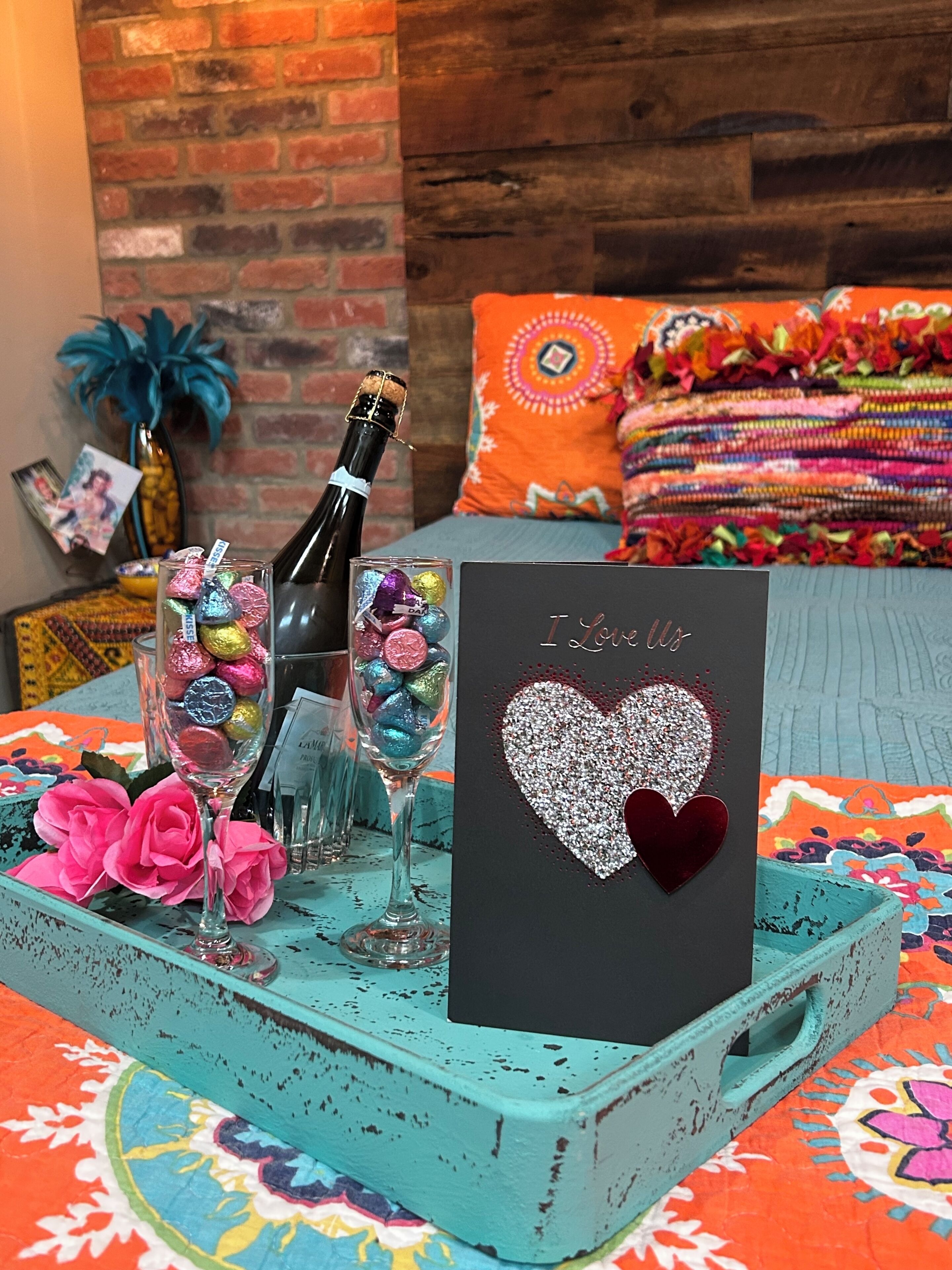 A teal tray with a bottle of champagne, colorful wrapped chocolates, pink roses, and a card with a glittery heart design.