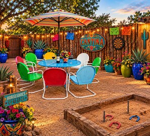 Un espacio de patio con cerramiento de madera, sillas coloridas, una mesa con sombrilla a rayas y un letrero que dice 'CANTINA'.