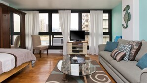 A modern living room with a glass coffee table, a sofa, a TV, and a view of buildings through large windows. - Oasis in Waikiki w/ Pool ⛱-Steps to the Beach, Zoo, SunDeck Waikiki Grand Hotel® (Honolulu)