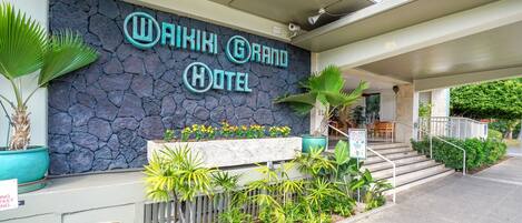 The exterior of the Waikiki Grand Hotel with a stone facade, outdoor seating area, and a sign indicating a full bar.