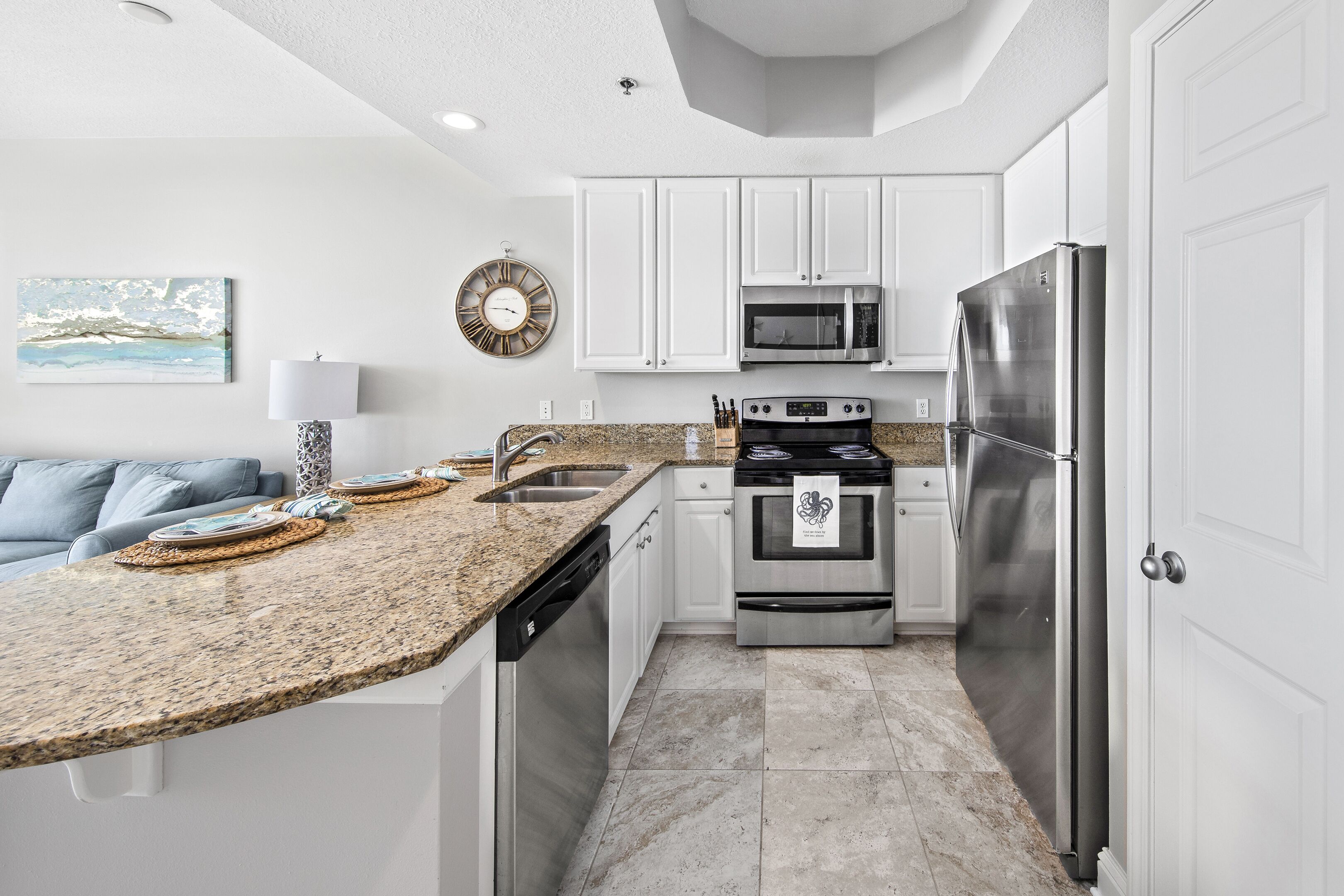 A modern kitchen with white cabinets, stainless steel appliances, a granite countertop, and a dining area with a blue couch.