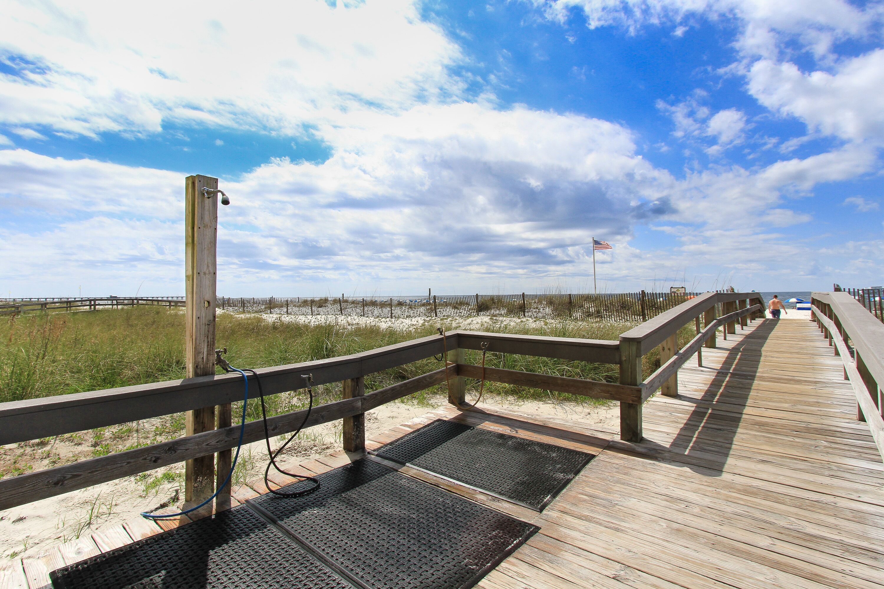 Une promenade en bois, dotée de grilles métalliques, qui mène à une plage de sable et d’herbe.