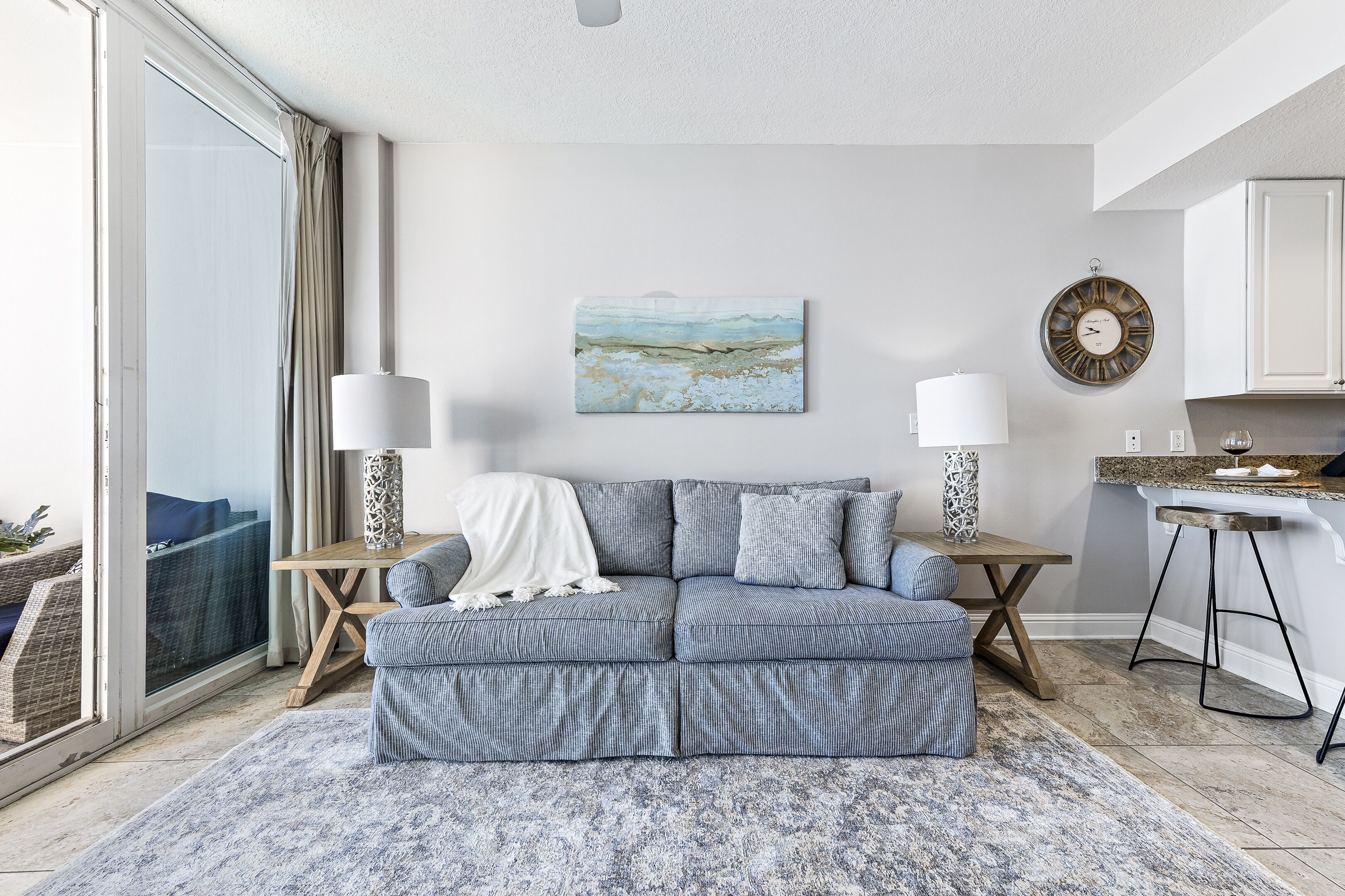 Una sala de estar moderna con un sofá gris, un cuadro, un reloj de pared redondo y una zona de cocina con un taburete de bar.