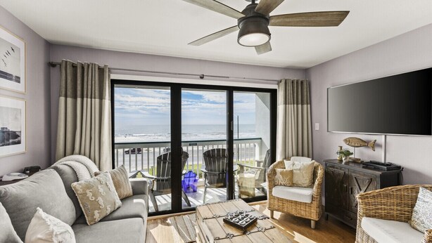Een woonkamer met een bank, stoelen, een salontafel, een televisie en een balkon met uitzicht op de oceaan.