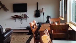 Uma sala de estar com teto de madeira, uma área de jantar com mesa e cadeiras, uma guitarra, uma televisão e uma janela com persianas.