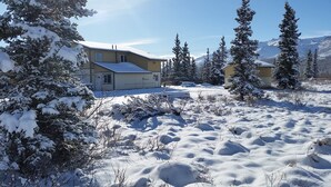 Paisagem coberta de neve com duas casas, uma delas com garagem, cercada por árvores carregadas de neve e montanhas ao fundo.