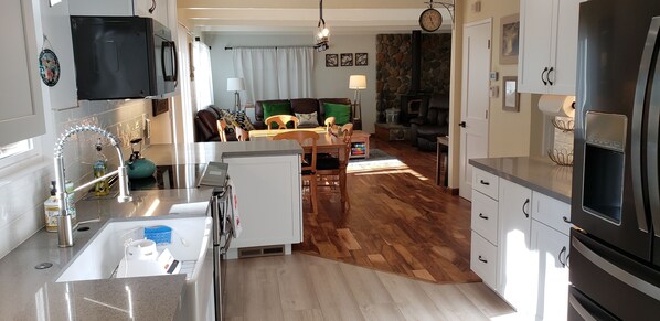 A modern kitchen with a built-in oven, a microwave, and a stainless steel refrigerator. There is a dining area with a table and chairs, and a living room with a sofa and a fireplace.