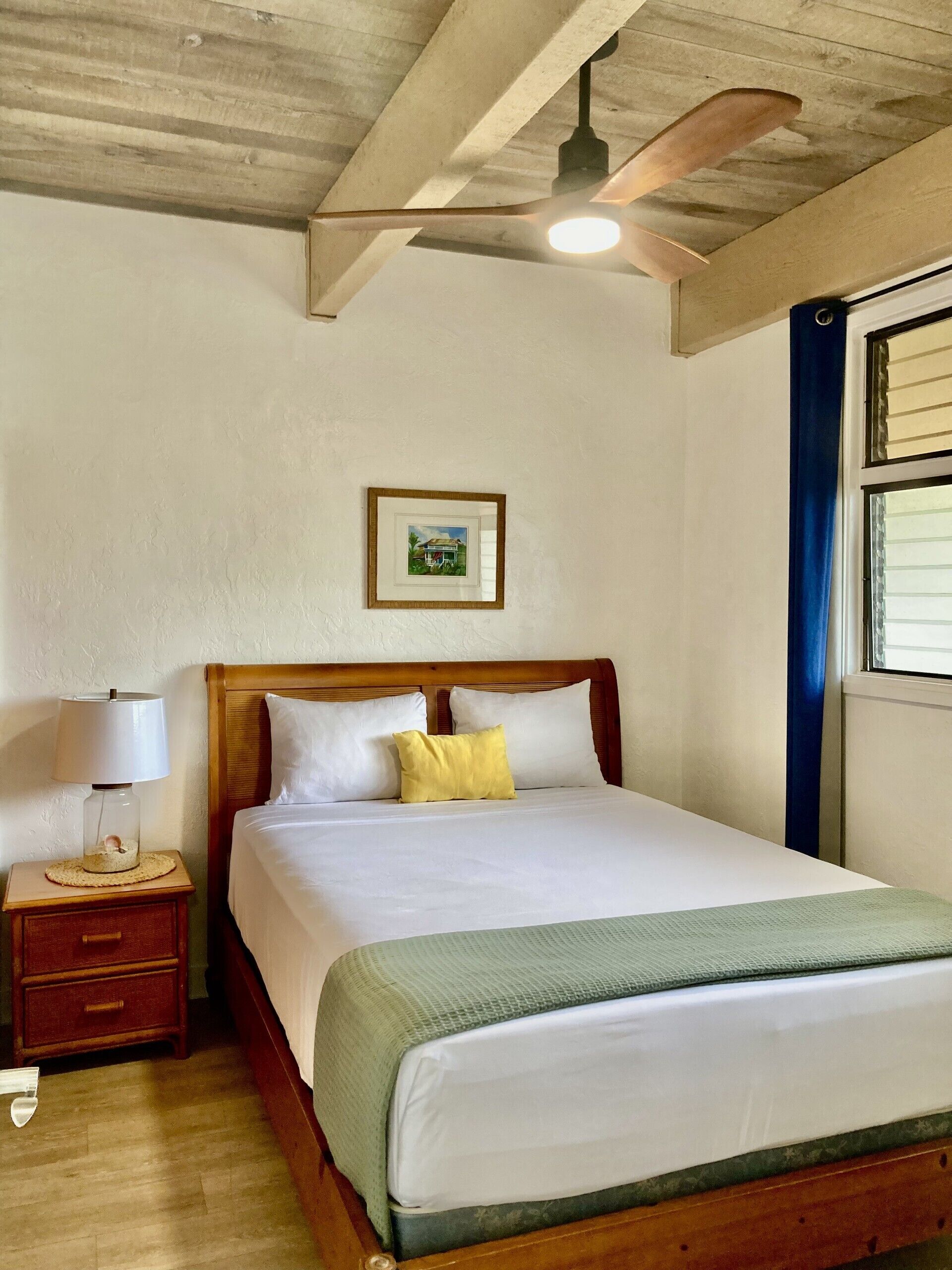 Una cama bien hecha con almohadas blancas y amarillas, un cabecero de madera, una mesita de noche con una lámpara y un cuadro en la pared.