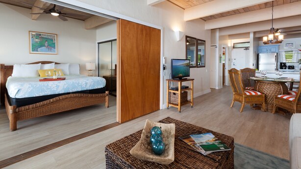 Una habitación de hotel con cama, televisor, zona de comedor con sillas y cocina visible a través de una puerta abierta.