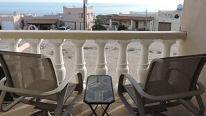 Un balcon avec deux chaises et une petite table, d’où l’on peut admirer une plage de sable et des bâtiments au loin.