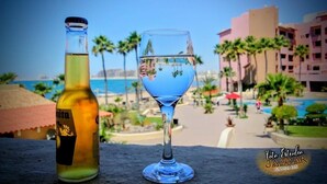 Une bouteille de bière Amita et un verre bleu, posés sur une étagère, avec vue sur une plage et des palmiers.