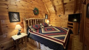 A cozy log cabin bedroom with a bed, a nightstand, a lamp, a television, and a framed picture on the wall. - Breathtaking views from decks! Private acres next door to Dollywood! Beautiful! (Pigeon Forge)