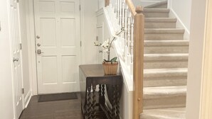 Een gang met een houten vloer, een witte deur, een trap met houten leuning en een aan de muur bevestigd hertenhoofd.