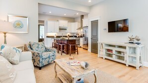 A spacious living room with a sofa, coffee table, and a TV. A kitchen is visible in the background with white cabinets and a refrigerator. - 4BR townhome near the beach with parking, WiFi, and pet-friendly stays. (Virginia Beach)