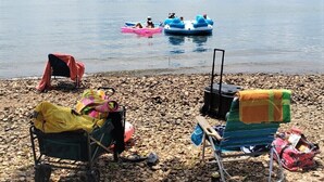 A pebbled beach with two beach chairs and a covered chair, a small cart, and a cooler. In the background, there are two inflatable boats on the water. - Newly Renovated Lakeside Cottage, Beautiful Lake Views With Adjacent Boat Ramp. (Dover)