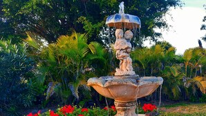 A stone fountain with water spouting from multiple levels, surrounded by lush greenery and red flowers. - SANIBEL SUN CASTLE  Splendid home, heated pool, great location, bike to beach. (Sanibel)