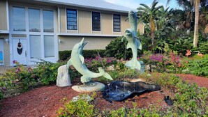 A building with a beige facade and a metal roof, surrounded by a landscaped garden with sculptures and tropical plants. - SANIBEL SUN CASTLE  Splendid home, heated pool, great location, bike to beach. (Sanibel)