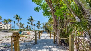 A pathway lined with rope barriers and palm trees leading to a beach area with buildings in the background. - Rare Walkout Condo On Sanibel Beach (Sanibel)