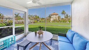 A room with a large window, a ceiling fan, a round table with a plant, a blue sofa, and a small bench. - Rare Walkout Condo On Sanibel Beach (Sanibel)