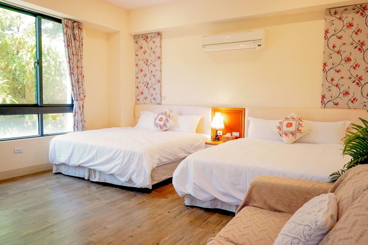 A hotel room with two beds, a wooden floor, a window with curtains, an air conditioner, and floral wallpaper.