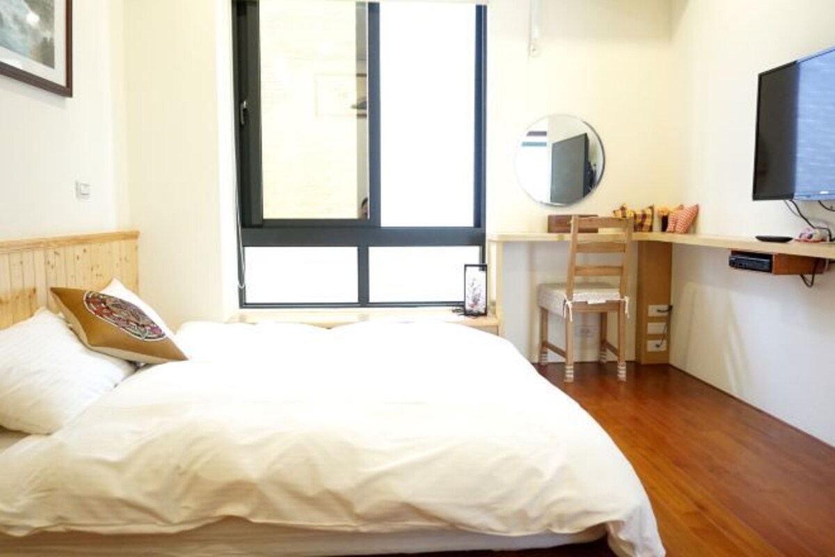 A bedroom with a bed, a desk, a chair, a TV, and a window.