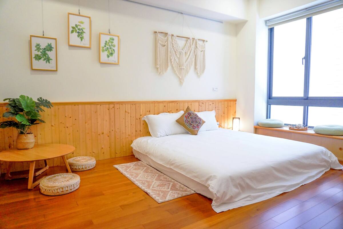 A bedroom with a wooden headboard, a bed with white bedding, a round wooden table with a plant, and a window with a view.