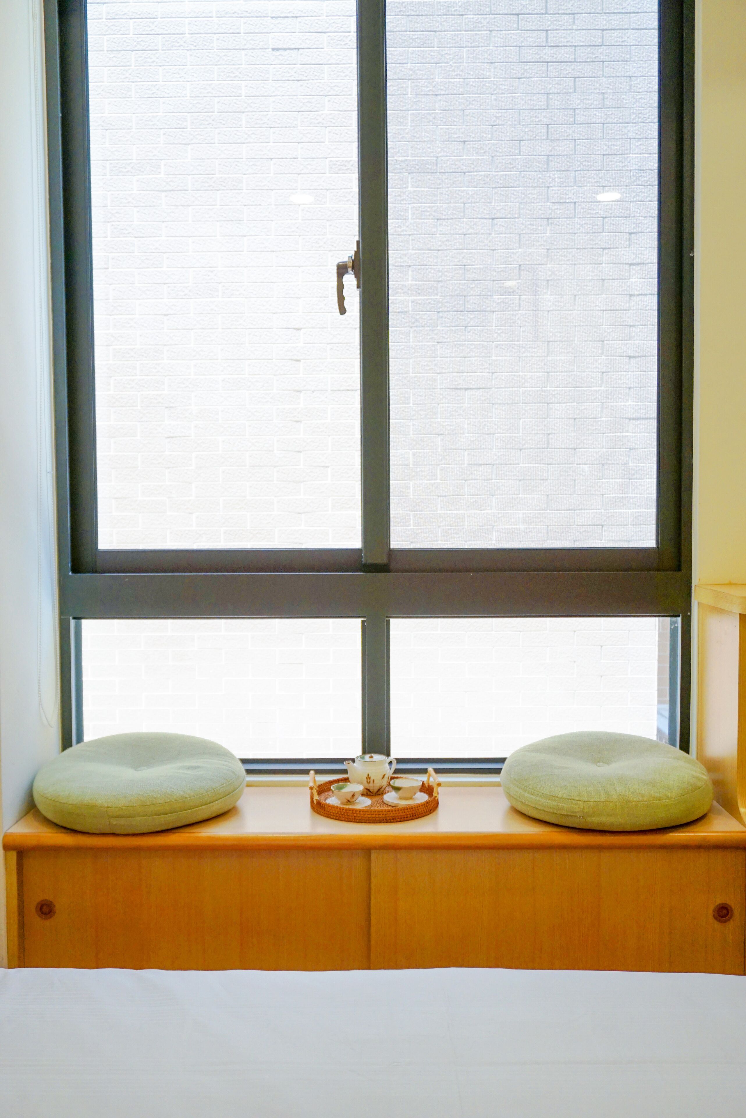 A window with a view of a brick wall, a wooden bench with two cushions, and a tray with a teapot and cups.