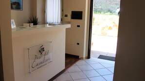 Un espace d’accueil avec une enseigne indiquant « Passo del Cavalier Bed & Breakfast », un comptoir de réception, une plante en pot, une fenêtre avec des rideaux et une vue sur un espace extérieur doté d’un mur en pierre.
