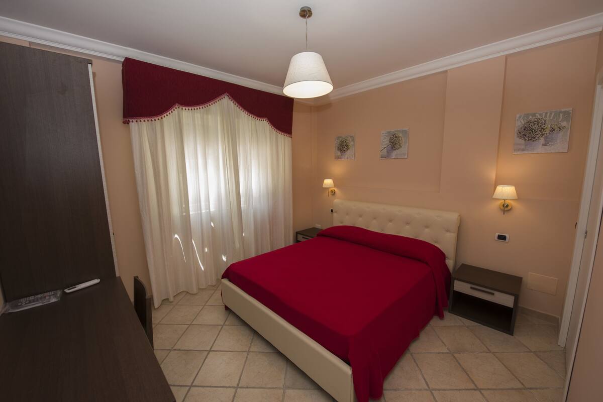 Un dormitorio con cama, mesitas de noche, una lámpara y un ventanal con cortinas.