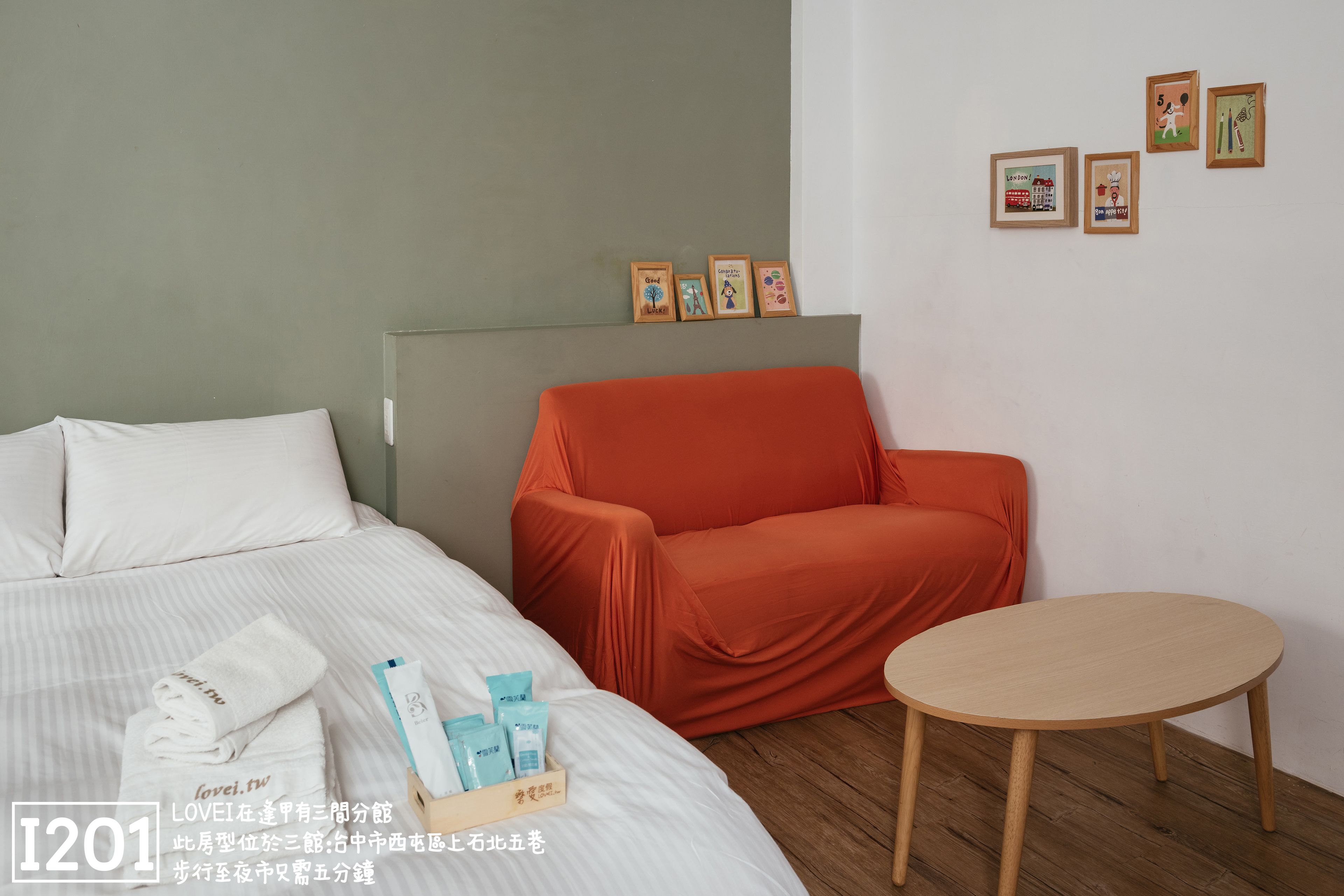 Una habitación de hotel con una cama, un sillón naranja tapizado, una mesita redonda y una pared con cuadros.
