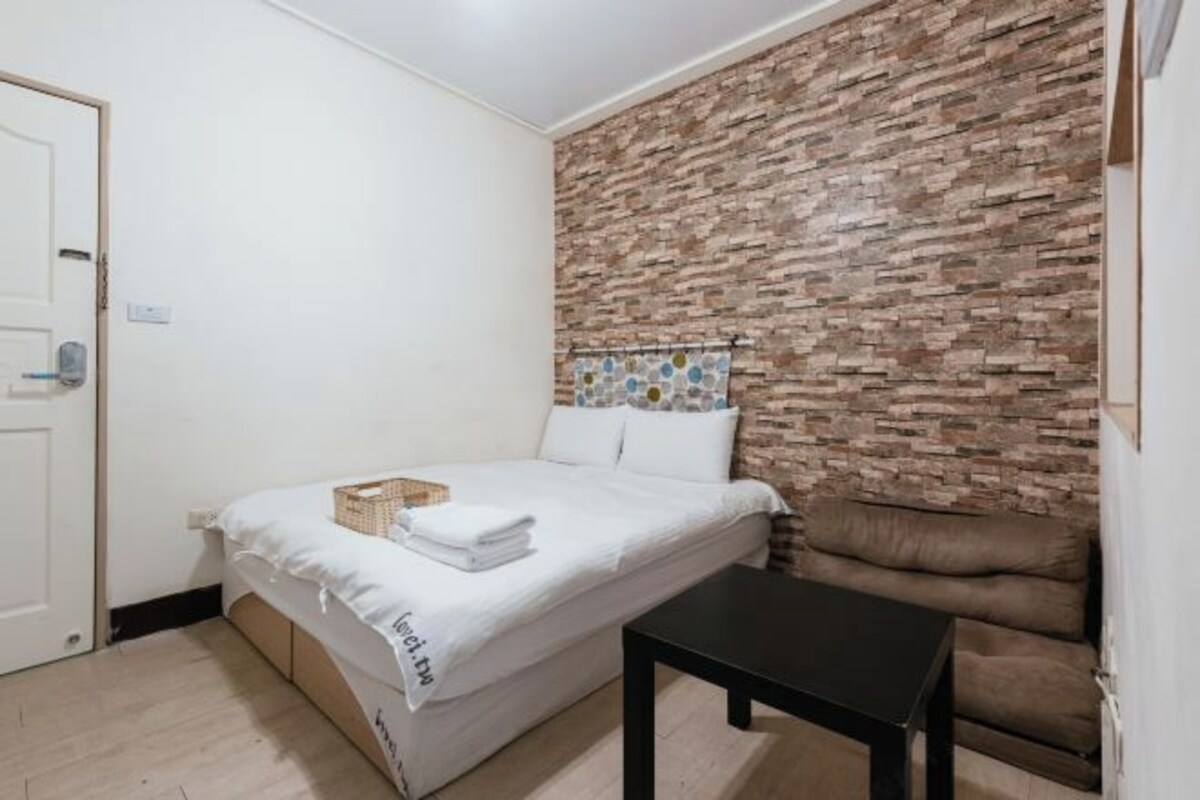 A bedroom with a bed, a small table, a brown sofa, and a brick-patterned wall.