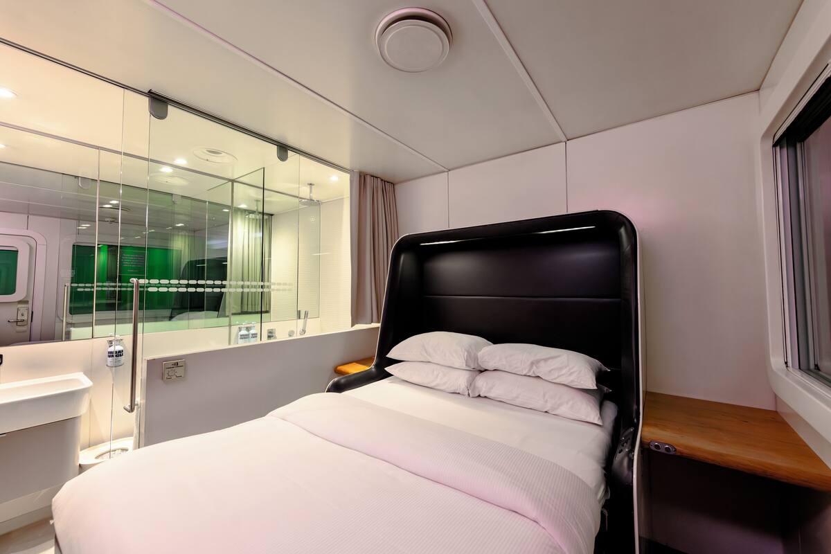 A modern hotel room with a bed, bedside table, and a glass partition separating the room from a bathroom.